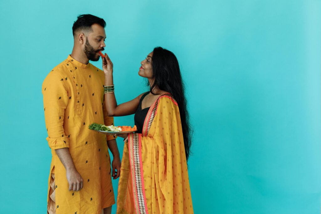 An Indian couple in traditional yellow attire shares a playful moment with food against a blue background.